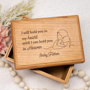 May include: A wooden keepsake box with a light brown finish. The lid features engraved text that reads "I will hold you in my heart until I can hold you in Heaven" and a line drawing of a mother and child. The name "Baby Fisher" is also engraved.