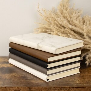 May include: A stack of five notebooks with different covers. The top notebook has a white marble pattern, the second is brown leather, the third is black leather, the fourth is grey, and the bottom is white.