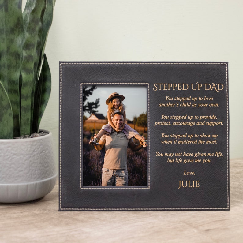 Peut inclure: Un cadre photo en cuir noir avec une inscription dor&eacute;e qui dit "Stepped Up Dad". L'inscription comprend un po&egrave;me sur un p&egrave;re qui s'engage &agrave; aimer et &agrave; soutenir un enfant. Le cadre comprend une photo d'un homme et d'une jeune fille.