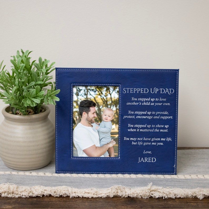 Peut inclure: Un cadre photo en cuir bleu avec une bordure cousue. Le cadre contient une photo d'un homme tenant un jeune enfant. Le cadre porte le texte "Stepped Up Dad" et un po&egrave;me sur le fait d'&ecirc;tre un beau-parent.