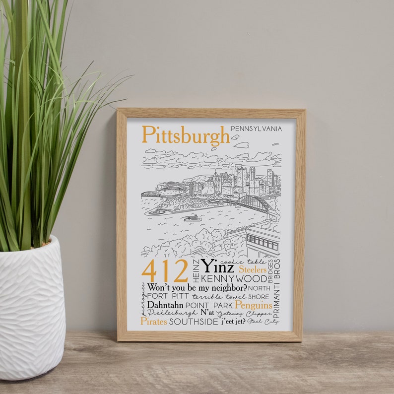 May include: A framed print with a black and white illustration of the Pittsburgh skyline. The print features the text "Pittsburgh, Pennsylvania" and a list of words and phrases associated with the city, including "412", "Yinz", "Kennywood", "Steelers", "Primanti Bros", "Won't you be my neighbor?", "North Shore", "Fort Pitt", "terrible towel", "Dahntahn", "Point Park", "Penguins", "Picklesburgh", "N'at", "Gateway Clipper", "Pirates", "Southside", "j'eet jet?", and "Steel City".