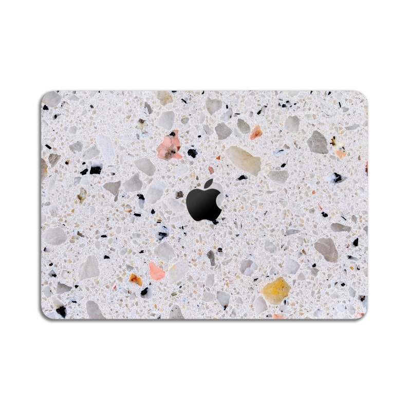 Puede incluir: Una piel de port&aacute;til blanca con un patr&oacute;n de terrazo. La piel tiene peque&ntilde;as motas negras, blancas, marrones y rosas por todas partes. El logotipo de Apple es visible en el centro de la piel.