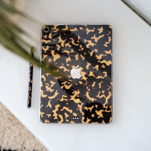 Può includere: Un iPad Apple con una custodia con motivo tartarugato e uno stilo abbinato. La custodia presenta un design nero e ambra con il logo Apple. Lo stilo è anch'esso decorato con lo stesso motivo. Gli articoli sono su una superficie bianca.