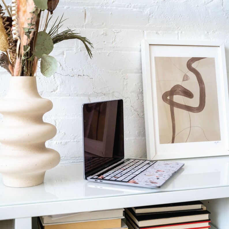 Puede incluir: Un ordenador port&aacute;til con una piel de teclado de m&aacute;rmol blanco y marr&oacute;n sobre una mesa blanca con un jarr&oacute;n de flores secas y una impresi&oacute;n de arte abstracto enmarcada en la pared detr&aacute;s.