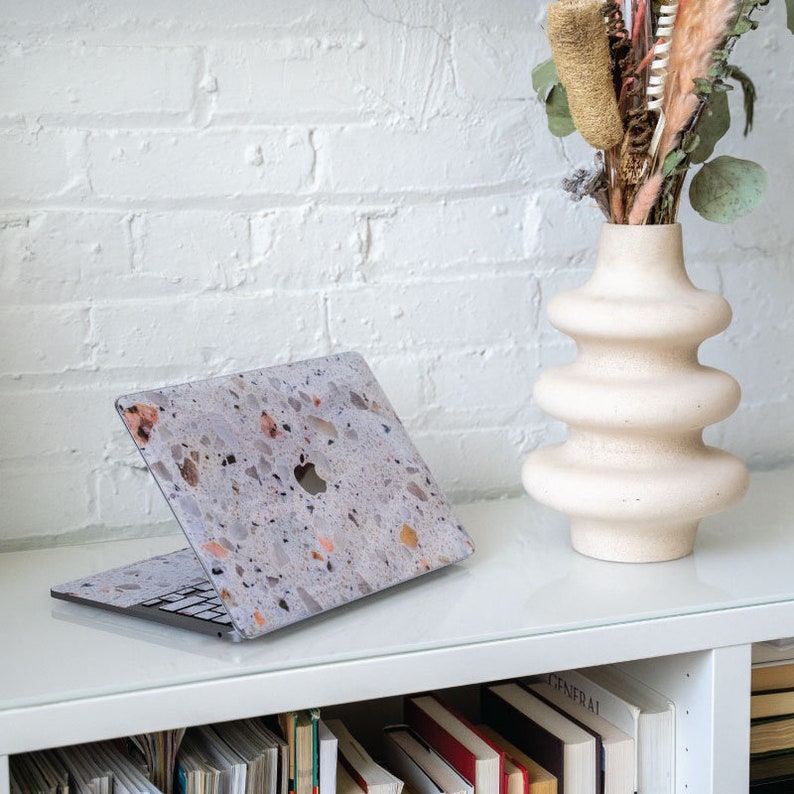 Puede incluir: Un ordenador port&aacute;til con una piel con estampado de terrazo en un estante blanco con libros. Un jarr&oacute;n con flores secas se encuentra en el estante a la derecha del port&aacute;til.