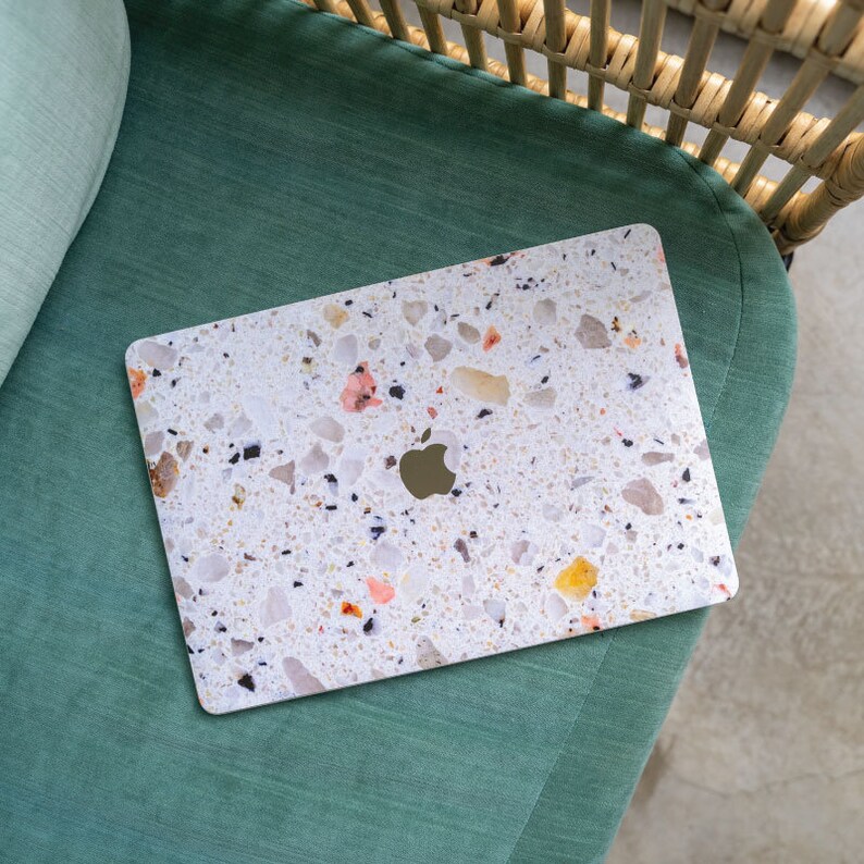 Puede incluir: Un ordenador port&aacute;til con un patr&oacute;n de terrazo blanco y beige en la tapa. El port&aacute;til est&aacute; sentado en una silla verde.