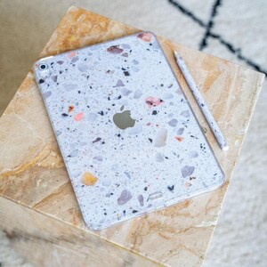 Op de afbeelding: Een Apple iPad met een terrazzo-patroon hoesje en bijpassende stylus. De hoes heeft een witte achtergrond met roze, zwarte en gele vlekken. De iPad ligt op een marmerachtig oppervlak.