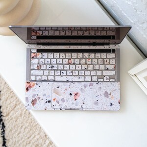 Puede incluir: Un ordenador port&aacute;til con una piel con estampado de terrazo que cubre el teclado, el trackpad y el exterior. La piel es de color beige claro con motas negras, grises y rosas.