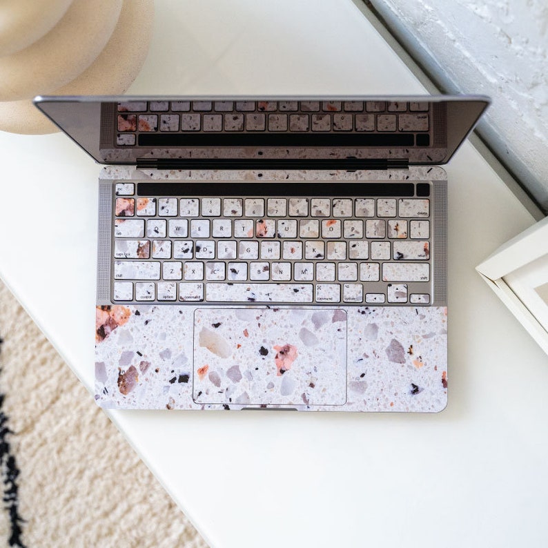Puede incluir: Un ordenador port&aacute;til con una piel con estampado de terrazo que cubre el teclado, el trackpad y el exterior. La piel es de color beige claro con motas negras, grises y rosas.