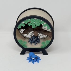 May include: A circular black frame display case featuring a tan interior with a brown tree branch, green leaves, and a white spider web. A small gray spider figure sits inside, while a bright blue spider figure rests in front of the display.