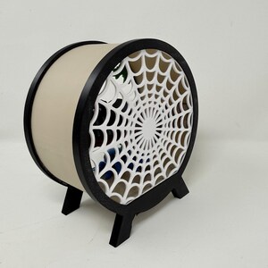 May include: A circular, drum-shaped enclosure featuring a black outer frame and a white, intricate spiderweb-patterned front panel. The structure is supported by two black feet and has a solid, light-colored cylindrical body with an open interior.