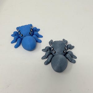 May include: Two small, 3D-printed spider figurines, one in bright blue and one in dark gray. Each spider features a rounded abdomen, eight articulated legs, and a head with large, cartoon-style eyes with white highlights on a black background.
