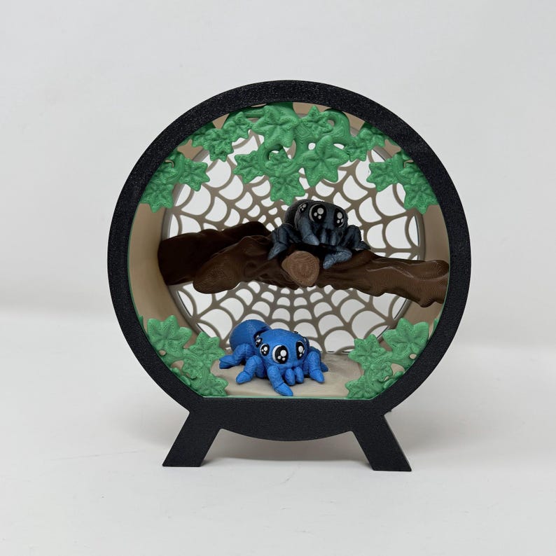 May include: A circular black frame containing a layered diorama featuring a brown branch, a web background, and green leaves. A black spider with large eyes sits on the branch, while a blue spider with large eyes rests at the bottom.