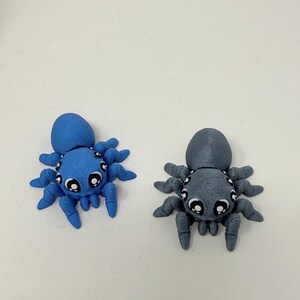 May include: Two small spider figurines, one bright blue and one gray, each featuring a rounded body, eight articulated legs, and large, expressive white and black eyes. The figures have a textured surface finish and are positioned side by side.