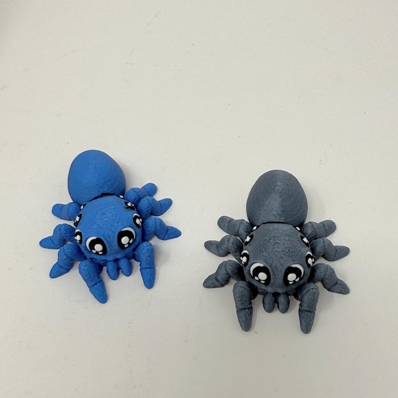 May include: Two small spider figurines, one bright blue and one gray, each featuring a rounded body, eight articulated legs, and large, expressive white and black eyes. The figures have a textured surface finish and are positioned side by side.