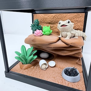 May include: A 3D printed terrarium display featuring a light-colored bearded dragon figurine perched on brown rock formations. The scene includes a green succulent, a pink flower, a white egg, a small hatchling, and a gray bowl filled with black insects.