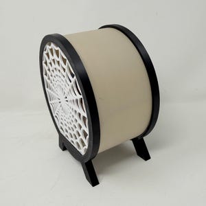 May include: A cylindrical object with a beige body and black circular frames on each side. One side features a white, intricate spider web pattern cutout. The structure is supported by two small black feet at the base, elevating the cylindrical form.