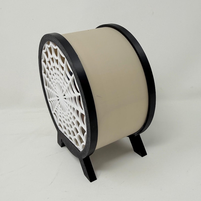 May include: A cylindrical object with a beige body and black circular frames on each side. One side features a white, intricate spider web pattern cutout. The structure is supported by two small black feet at the base, elevating the cylindrical form.