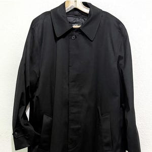 May include: A solid black trench coat featuring a pointed collar, a concealed button placket, long sleeves with buttoned cuffs, and two side pockets. The coat has a smooth, structured finish and hangs straight, providing a clean, classic silhouette.