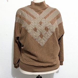 Op de afbeelding: Een bruine trui met lange mouwen en een opstaande kraag, voorzien van een textuurstof en een contrasterend beige en bruin pied-de-poulepatroon op de borst en schouders. Het patroongedeelte is versierd met kleine, gelijkmatig verdeelde goudkleurige studs.