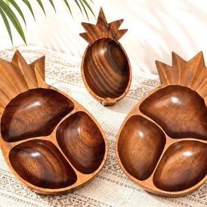 May include: Three pineapple-shaped wooden bowls with a dark brown finish and visible wood grain. Two of the bowls are divided into three sections, while the smaller central bowl is a single piece. Each bowl features a carved leaf top design.
