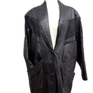 May include: A black leather coat featuring a shawl collar, long sleeves, and two front buttons. The garment includes textured black fabric panels on the upper chest and decorative stitching details on the two front pockets, creating a structured look.