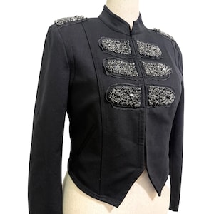May include: A black military-style jacket featuring a high collar, long sleeves, and three horizontal rows of dark, faceted crystal embellishments on the chest. The shoulders are accented with matching dark crystal epaulets.