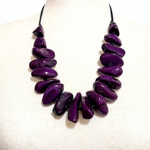 May include: A statement necklace featuring a series of smooth, polished, deep purple, irregular pebble-shaped beads strung together on a thin black cord. The beads are arranged in a graduated pattern, creating a bold, chunky, and vibrant accessory.