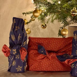 以下が含まれることがあります： クリスマスをテーマにしたギフトラッピング。 金色の雪の結晶が付いた濃い青色の生地と、金色の星模様の赤い生地が装飾的な結び目で結ばれています。 金色のオーナメントが背景のクリスマスツリーから吊り下げられています。