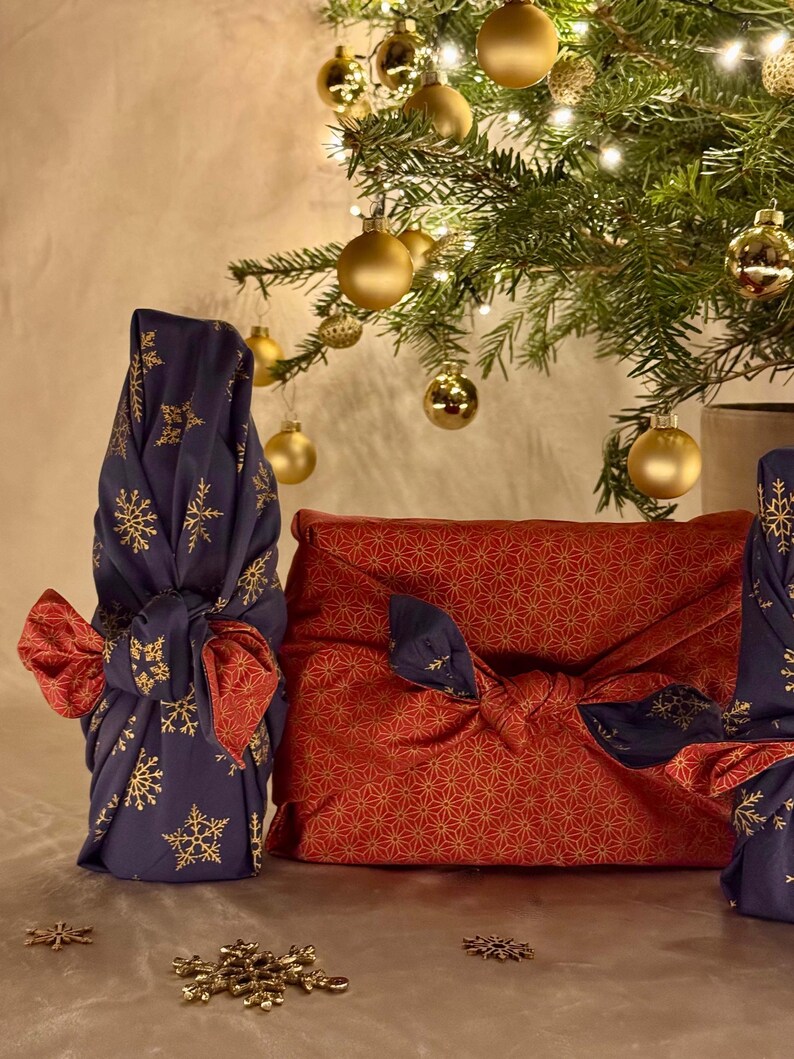 以下が含まれることがあります： クリスマスをテーマにしたギフトラッピング。 金色の雪の結晶が付いた濃い青色の生地と、金色の星模様の赤い生地が装飾的な結び目で結ばれています。 金色のオーナメントが背景のクリスマスツリーから吊り下げられています。