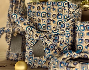 Può includere: Diverse scatole regalo rettangolari avvolte in tessuto con un motivo a ventaglio ripetuto in blu, oro e bianco. Il tessuto è annodato al centro di ogni pacchetto, con piccole etichette regalo rettangolari nere attaccate ai nodi di tessuto.