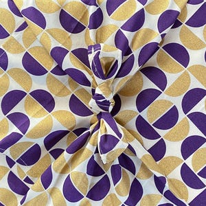 Op de afbeelding: Een textielstof met een herhalend geometrisch patroon van paarse en gouden halve cirkels op een witte achtergrond. Het materiaal is in het midden samengebonden tot een knoop, wat de symmetrische rangschikking van de vormen benadrukt.