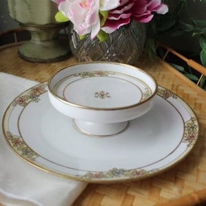 May include: A white porcelain pedestal serving dish with a gold rim and floral pattern. The dish is on a woven bamboo tray.