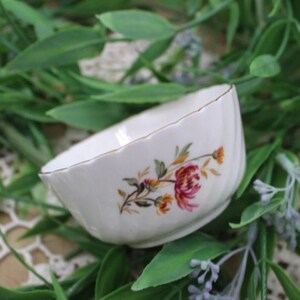 Op de afbeelding: Een kleine witte porseleinen kom met een gegolfde rand en een bloemenmotief. De kom heeft een roze en gele bloem met groene bladeren.