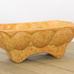 May include: A yellow ceramic planter with a speckled glaze and a unique, rounded design. The planter has four legs and is perfect for displaying small plants or succulents.