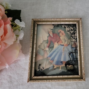 May include: A framed silhouette artwork depicting a couple in a garden setting. The man is wearing a top hat and the woman is wearing a long dress. The artwork is in a light brown frame with a decorative border.