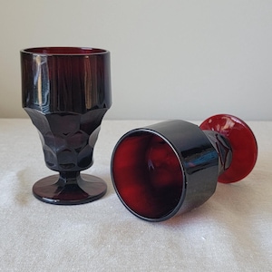 May include: Two dark red glass goblets with a textured, faceted design. The goblets have a round base and a wide, round bowl.