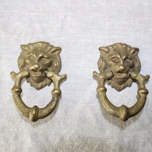 May include: Two antique brass lion head drawer pulls with a ring handle. The lion heads are detailed with a mane and a snarling expression.