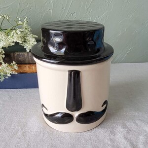 May include: A white ceramic jar with a black lid and a black mustache and nose design. The jar has a textured lid and a smooth body.