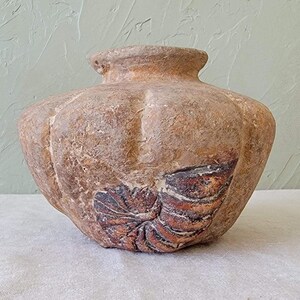 May include: A textured, brown ceramic vase with a rounded body and a narrow neck. The vase features a leaf-like design in shades of brown and red. The vase is displayed on a white surface against a light green background.