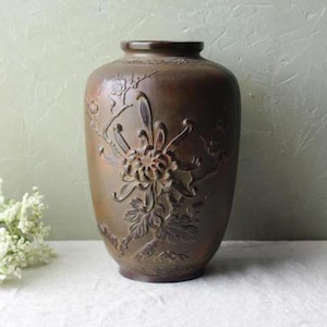 May include: A dark brown ceramic vase with an intricate design of a flower and leaves. The vase has a rounded body and a narrow neck.