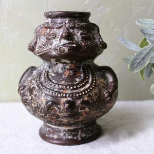 May include: A brown ceramic vase with a textured surface and intricate carvings. The vase is decorated with white and brown paint and has a unique, almost human-like face.