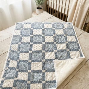 May include: A rectangular quilt featuring a patchwork design of alternating squares in dusty blue floral patterns and cream fabric with small blue sprigs. The quilt has a solid cream-colored backing and is finished with a white fabric binding.