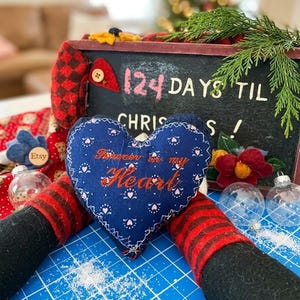 May include: A dark blue fabric heart ornament with a white floral pattern and the embroidered text "Forever in my Heart" in orange. The heart is positioned in front of a black chalkboard sign with the text "124 DAYS TIL CHRISTMAS!" in pink and white.