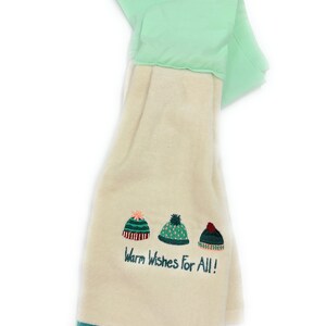 Pu&ograve; includere: Un asciugamano da cucina color crema con un bordo verde. Presenta tre cappelli invernali ricamati e il testo "Warm Wishes For All!"