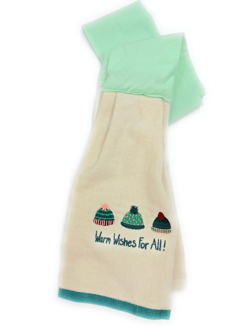Pu&ograve; includere: Un asciugamano da cucina color crema con un bordo verde. Presenta tre cappelli invernali ricamati e il testo "Warm Wishes For All!"