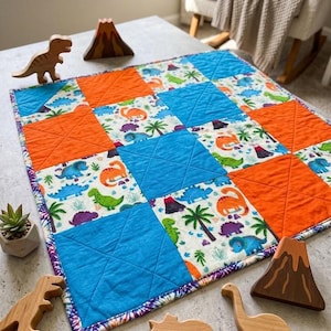 May include: A square patchwork quilt featuring alternating blocks of solid bright orange, solid blue, and a white fabric printed with colorful dinosaurs, palm trees, and volcanoes. The quilt is surrounded by wooden dinosaur and volcano figures.