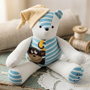 May include: A stuffed bear made from white and light blue striped fabric, wearing a yellow nightcap. The bear features a brown monkey appliqué on its chest with a yellow crescent moon and the embroidered names Alex & Ant on its foot.