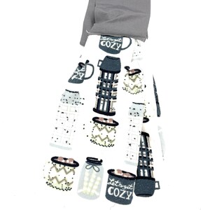 Pu&ograve; includere: Un asciugamano da cucina bianco con un motivo grigio e bianco di tazze e barattoli. Il testo "Let's get cozy" &egrave; stampato su una delle tazze. L'asciugamano &egrave; piegato e ha un anello di tessuto grigio nella parte superiore.