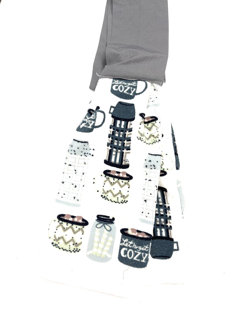 Pu&ograve; includere: Un asciugamano da cucina bianco con un motivo grigio e bianco di tazze e barattoli. Il testo "Let's get cozy" &egrave; stampato su una delle tazze. L'asciugamano &egrave; piegato e ha un anello di tessuto grigio nella parte superiore.